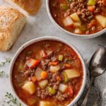 Two bowls of hearty hamburger soup with potatoes, carrots, and celery next to crusty bread